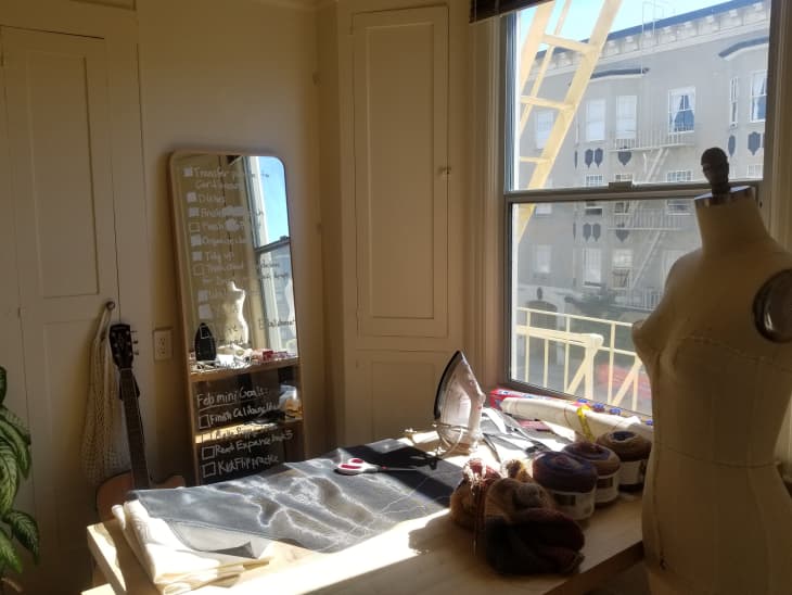 Sewing room with mannequin, fabric, yarn, iron, and a guitar by a window with a view of a building and fire escape.