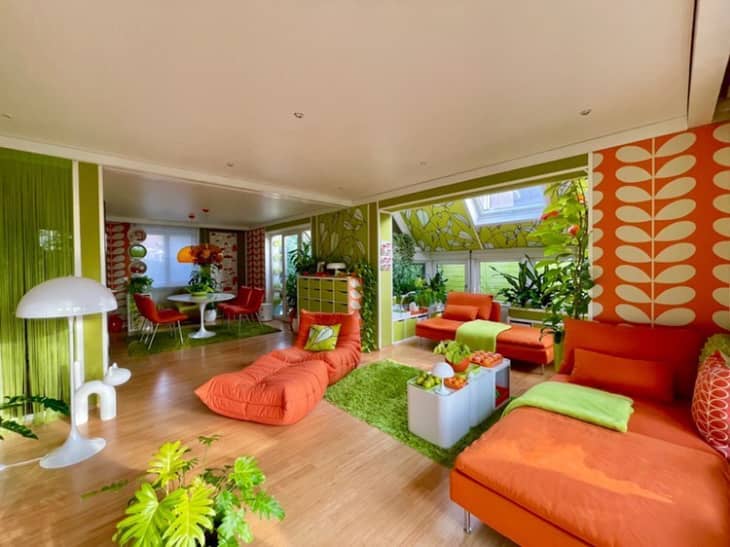 Retro living room with orange and green decor, patterned wallpaper, and modern furniture, featuring plants and a dining area.