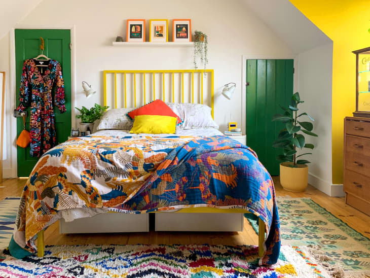 bedroom, yellow slated headboard, angled ceiling, green doors, plants, colorful comforter, multi-colored rug, wood floors, sunlight