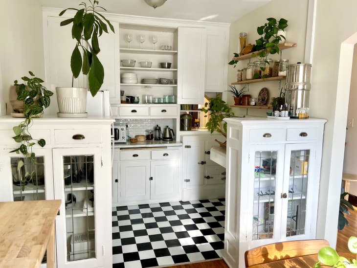 kitchen with white cabinets with black pulls, black and white checkered floor, narrow wood wall shelves with food storage jars and plants, small built in extra counter space, buff colored wall, white cabinet with glass doors, main kitchen cabinet open to show white shelves with glasses and dishes