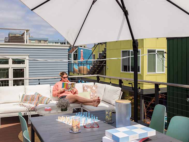 A woman reading a colorful book on a rooftop patio with a white sectional sofa, chess set, and vibrant buildings in the background.