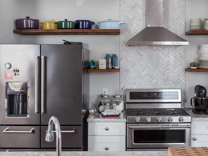 Modern kitchen featuring a stainless steel refrigerator, gas stove, and herringbone tile backsplash, with colorful cookware on shelves.