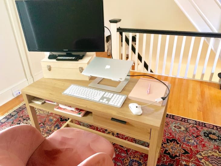 Home office setup with a wooden desk, laptop stand, keyboard, mouse, and a pink chair on a patterned rug.