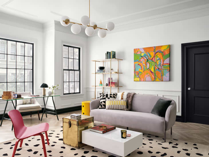 Modern living room with gray sofa, abstract art, glass desk, pink chair, and geometric rug.