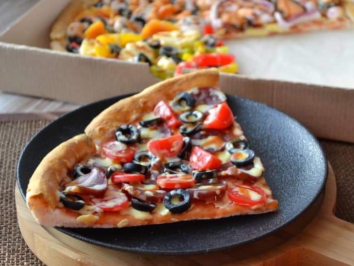 Pizza slice with tomatoes, olives, and cheese on a black plate, with a pizza box in the background.