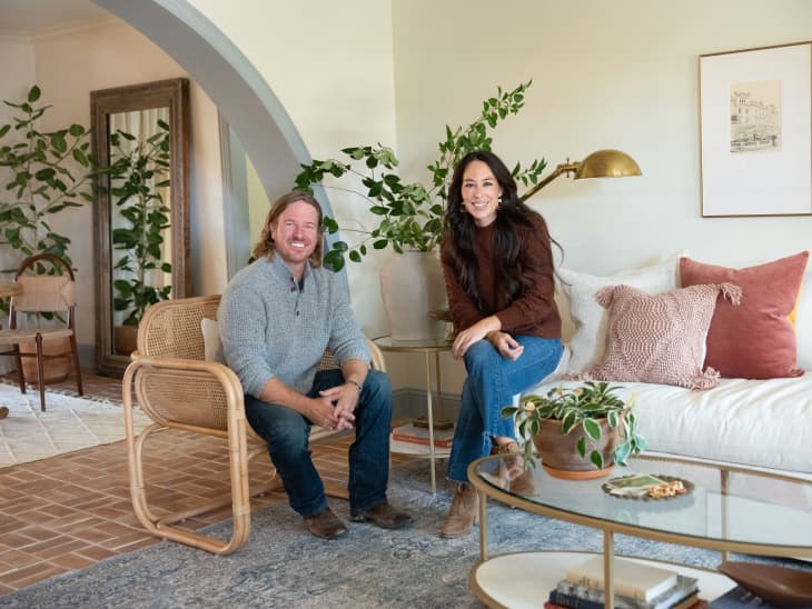 A couple sitting in a cozy living room with plants, a rattan chair, and a glass coffee table with decor items.