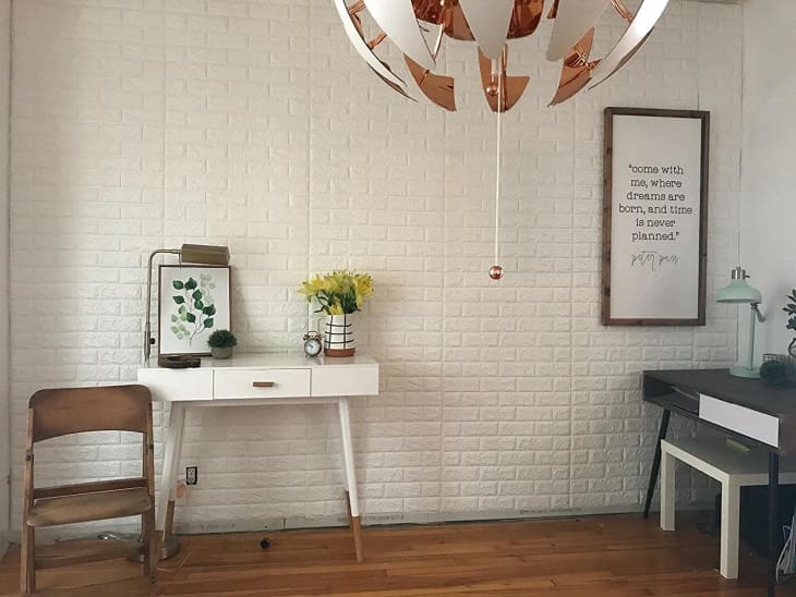 Home office with white desk, wooden chair, leafy artwork, yellow flowers, and a framed quote on a brick wall.