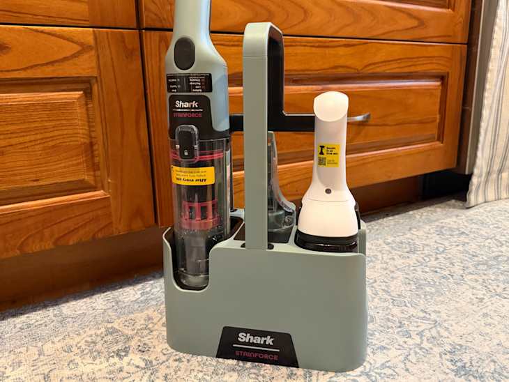 Shark Stainforce cordless vacuum and handheld cleaner in a storage dock, set against wooden cabinets and a patterned rug.
