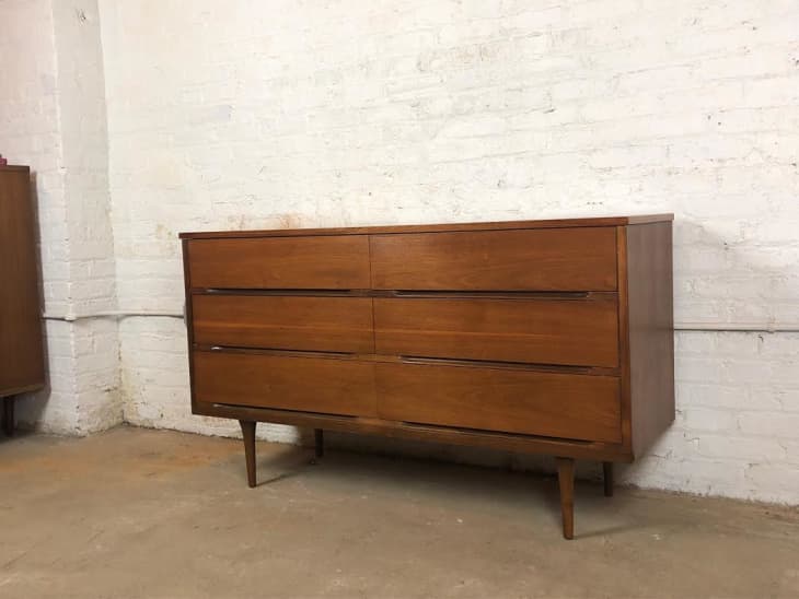 Mid-century modern wooden dresser with six drawers against a white brick wall.