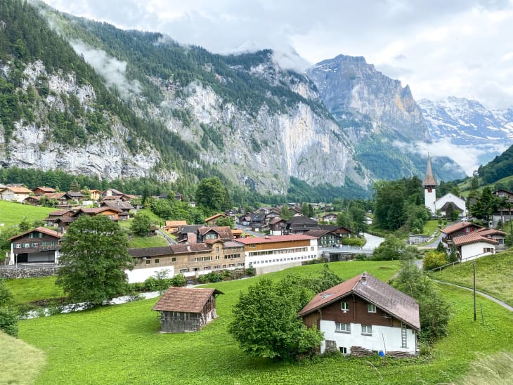 Village nestled in a lush valley with wooden houses, a church, and towering mountains in the background.