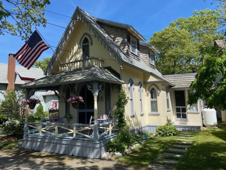 Charming yellow cottage with a porch, hanging flowers, and an American flag, surrounded by greenery.