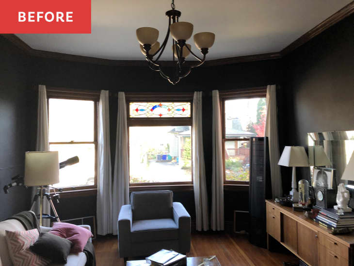 Living room with dark walls, bay windows, chandelier, telescope, blue armchair, and wooden sideboard with lamps.