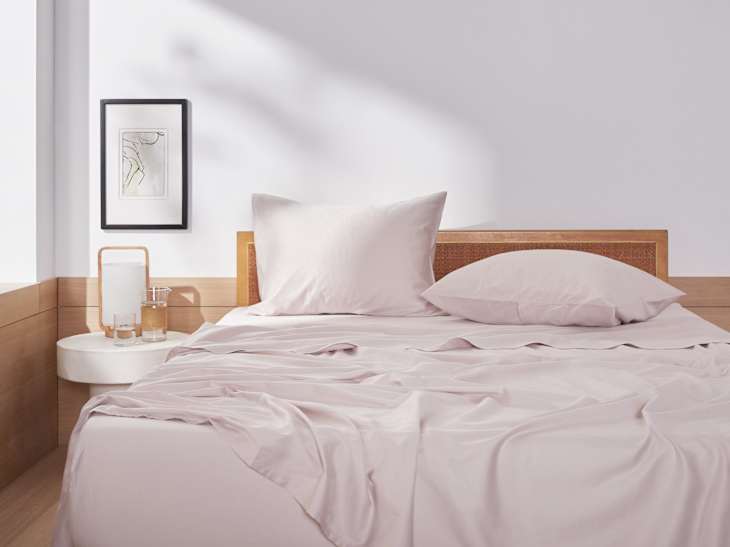 Minimalist bedroom with light pink bedding, wooden headboard, framed art, and a side table with a lamp and glass of water.