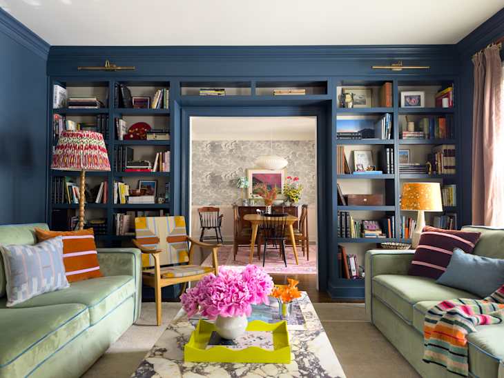 Cozy living room with green sofas, colorful pillows, a marble coffee table, and a bookshelf filled with books.