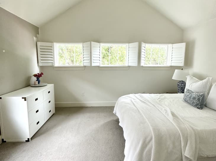 beige walls with a white 6 drawer dresser, white bed linens, beige carpet and white shutters on small horizontal windows