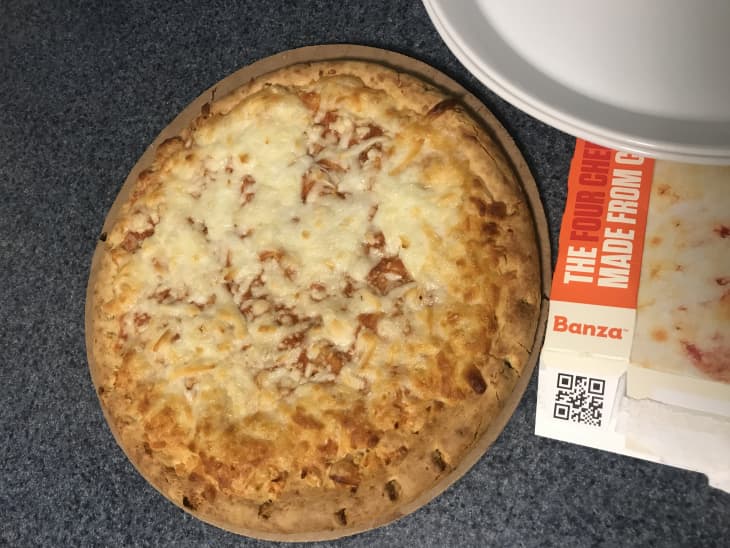 Cheese pizza on a countertop next to a Banza box with a white plate nearby.