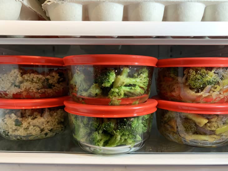 Glass containers with red lids filled with quinoa, broccoli, and vegetables in a refrigerator.