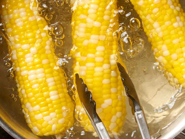 three ears of shucked corn in a boiling pot of water with tongs coming in
