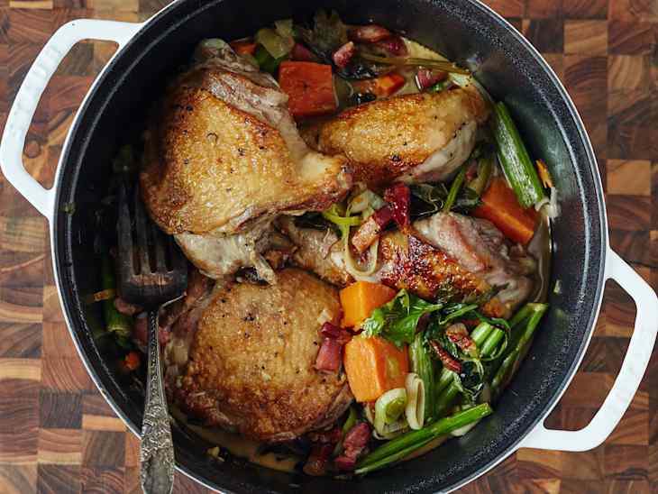 Braised chicken thighs with vegetables including carrots, greens, and bacon in a black pot on a wooden surface.