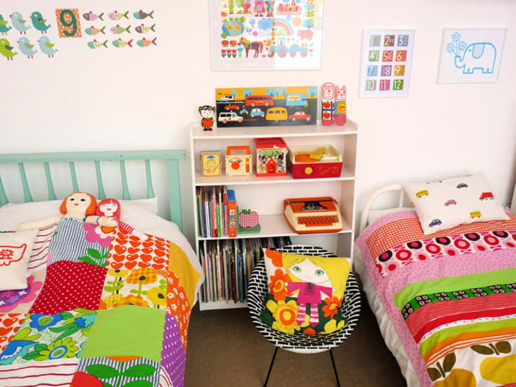 Colorful children's bedroom with two beds, vibrant quilts, a shelf with toys and books, and playful wall art.