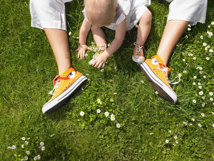 shot from above, a baby sitting inbetween someone's legs on the grass. grass has little white flowers. baby is wearing white clothes and orange hi-top sneakers. you only see the adult's legs and they are wearing orange hi-top sneakers as well.