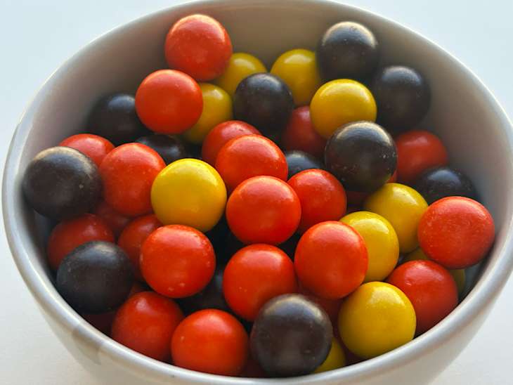 Colorful round candies in a white bowl, featuring red, yellow, black, and orange hues.