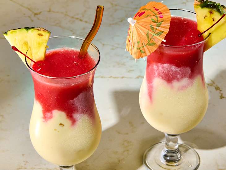Two tropical drinks with layered red and yellow slush, garnished with pineapple slices and colorful umbrellas.