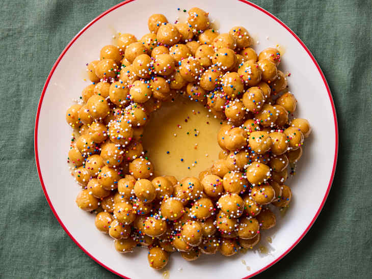 A circular arrangement of golden fried dough balls topped with colorful sprinkles and drizzled with syrup on a white plate.