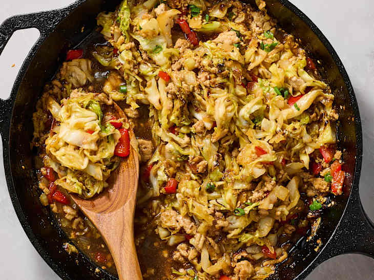 A cast iron skillet filled with stir-fried cabbage, ground meat, red bell peppers, and green onions, garnished with sesame seeds.