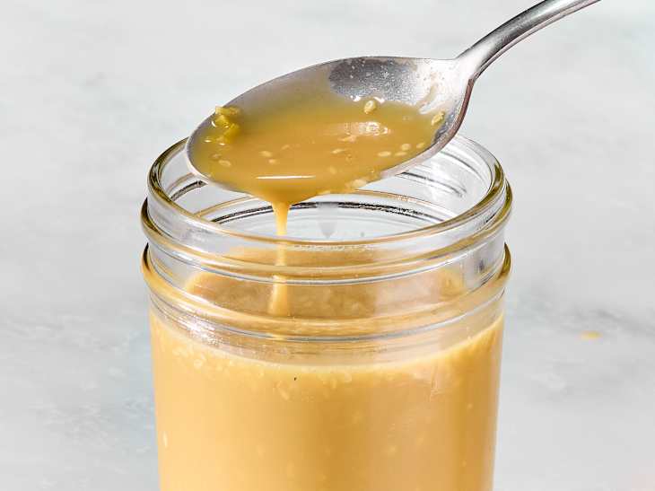 Creamy sauce dripping from a spoon into a glass jar, with a smooth texture and light brown color.
