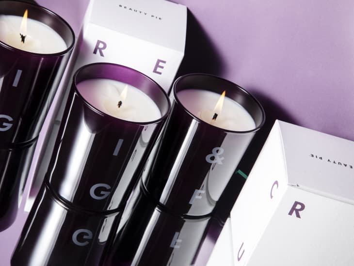 Three lit candles in dark glass holders with white boxes labeled "Beauty Pie" on a purple surface.