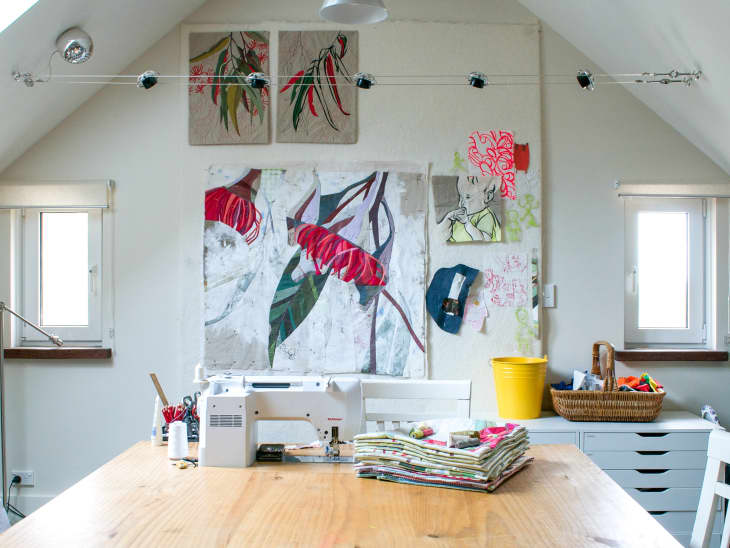 Sewing room with a wooden table, sewing machine, fabric stacks, and colorful artwork on the wall.