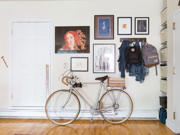 Bicycle against a wall with framed art, jackets, and backpacks hanging on hooks.