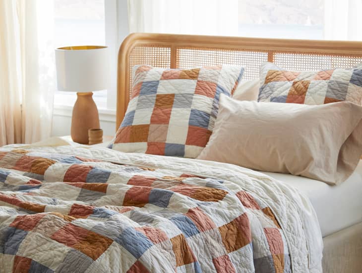 Patchwork quilt and pillows in blue, orange, and white on a bed with a wooden headboard, next to a ceramic lamp.