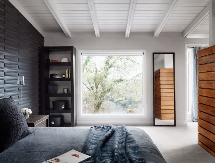 Modern bedroom with dark accent wall, large window, and wooden dresser. Gray bedding and a blue throw blanket on the bed.