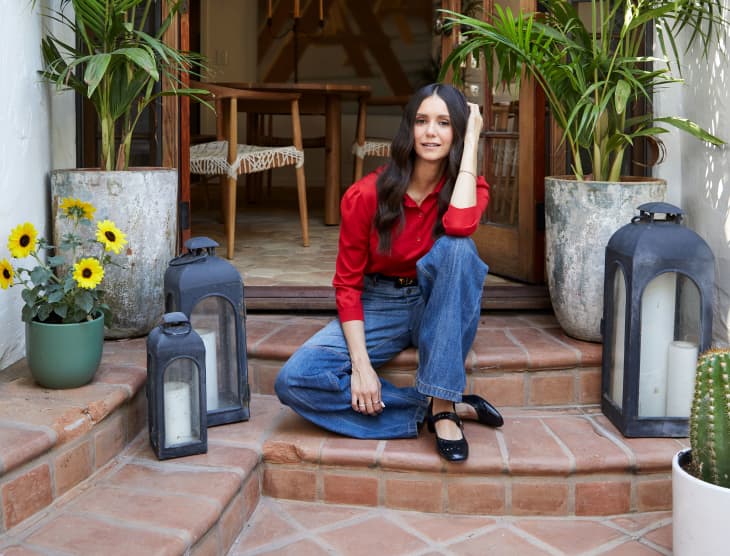 Woman in red shirt and jeans sitting on terracotta steps, surrounded by lanterns, sunflowers, and potted plants.