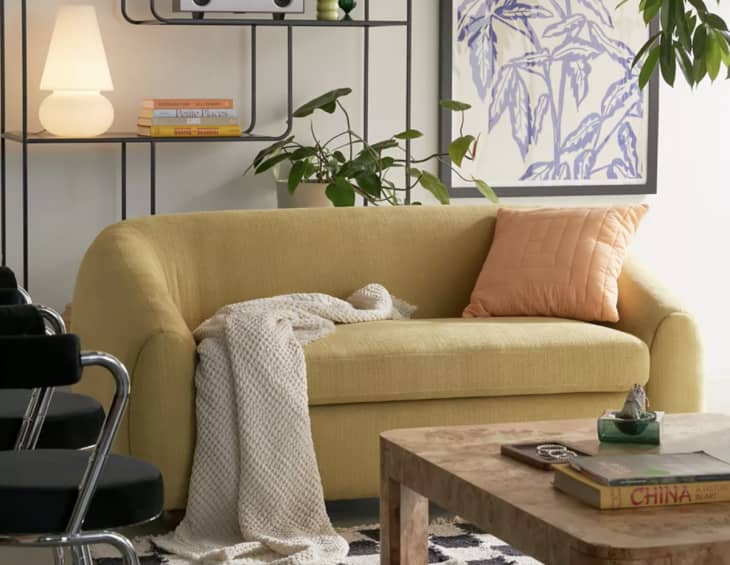 Yellow sofa with orange pillow, cream throw, and plant on shelf in cozy living room setting.
