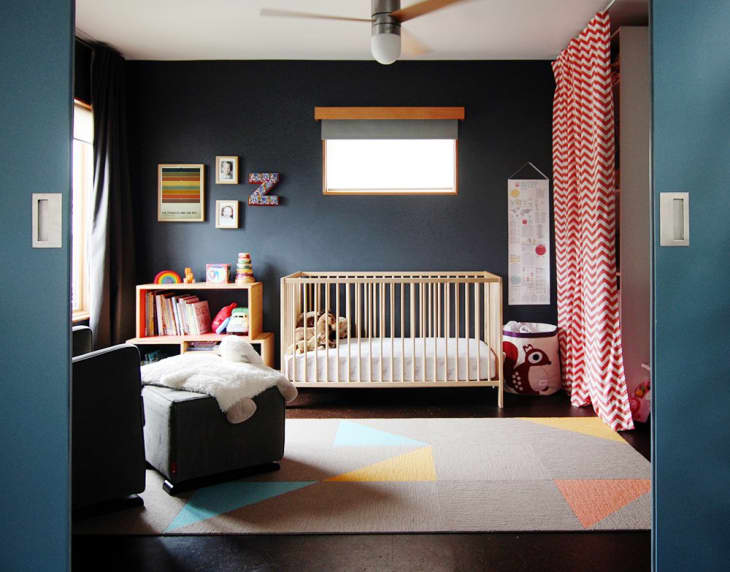 Nursery with a wooden crib, dark blue walls, colorful rug, and red chevron curtains, featuring books and toys on shelves.
