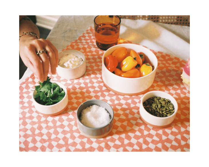 Hand reaching for cilantro on a patterned tablecloth with bowls of peppers, seeds, salt, and cream.