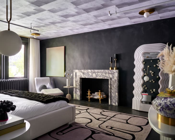 Modern bedroom with a marble fireplace, abstract rug, white chair, and decorative vases on a black wall.