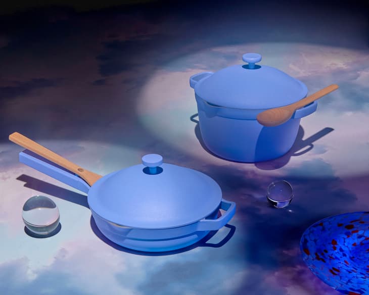 Blue cookware set with wooden handles, including a pot and pan, on a cloud-patterned surface with glass spheres.