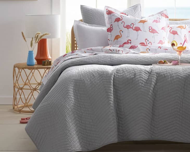 Gray quilted bedding with flamingo print pillows, wicker side table, orange lamp, and a cocktail glass.