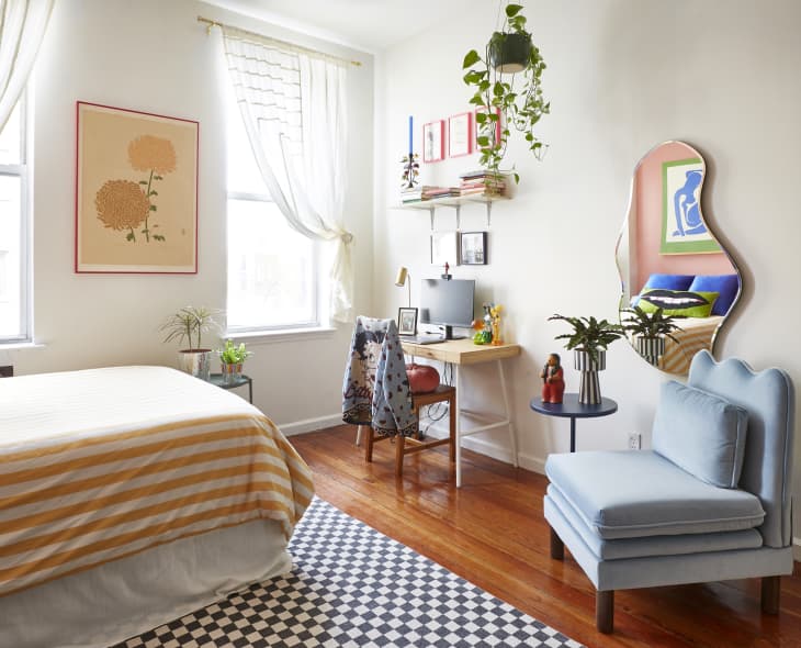 Bedroom with striped bedspread, blue chair, wavy mirror, desk with computer, hanging plant, and framed art on walls.