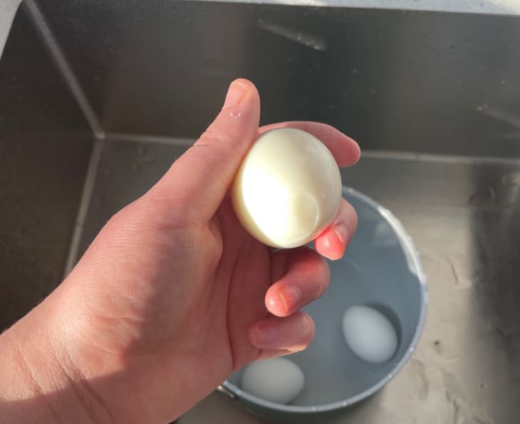 Hand holding a peeled hard-boiled egg over a sink with eggs in a bowl of water.
