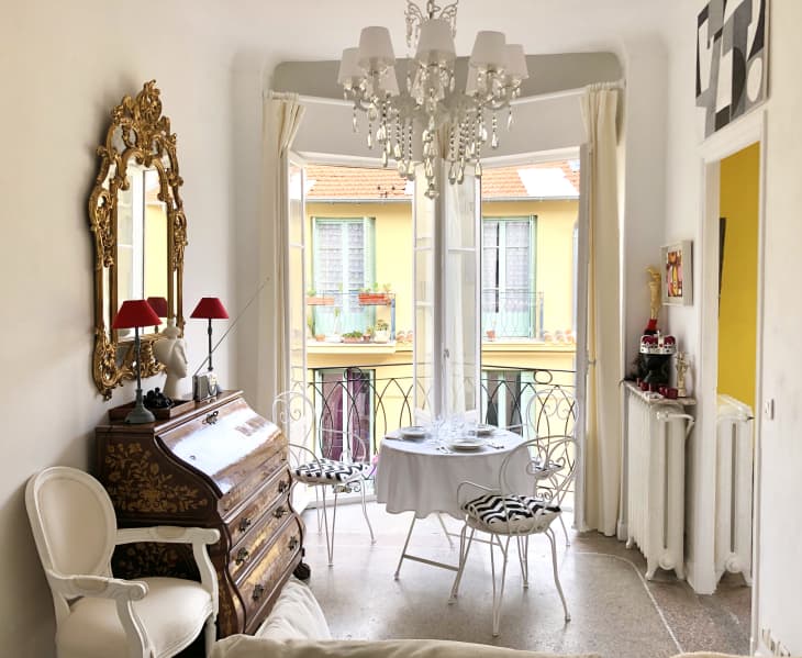 Elegant room with ornate mirror, chandelier, antique desk, and small dining table by a window with wrought iron chairs.