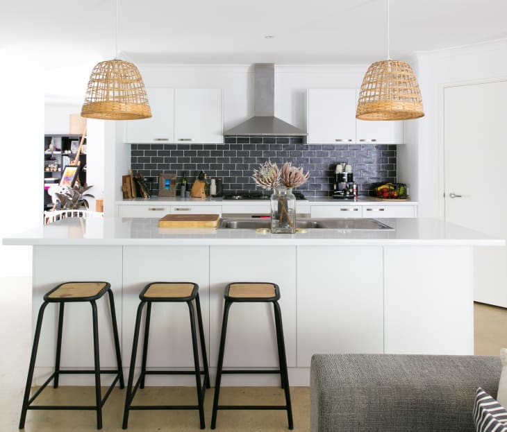 Modern kitchen with white island, black stools, wicker pendant lights, and black tile backsplash.