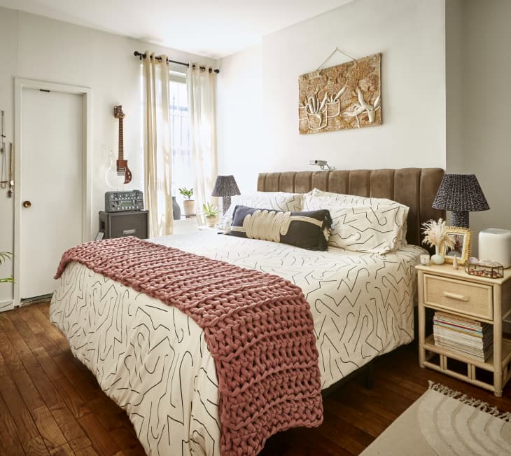 Cozy bedroom with a patterned bedspread, pink knit throw, guitar on wall, and bedside table with lamps and decor.