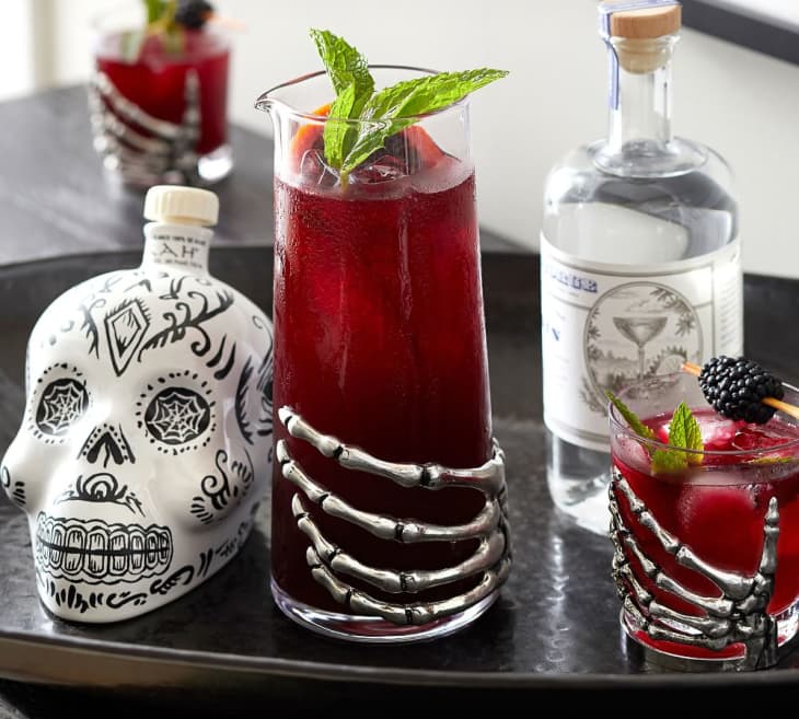 Skull-themed bottle with red cocktails in skeleton-hand glasses, garnished with mint and blackberries.