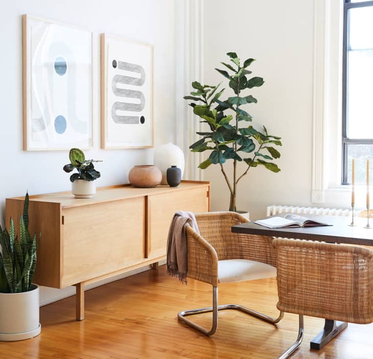 Mid-century modern dining room with wooden sideboard, abstract art, woven chairs, and potted plants.