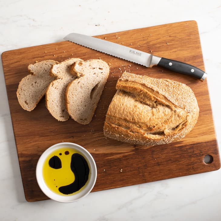 Sliced bread on a wooden cutting board with a serrated knife and a bowl of olive oil and balsamic vinegar.